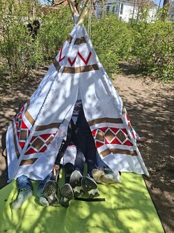 Das Bild zeigt ein Tipi-Zelt in einem Außenbereich. Kinder sitzen versteckt darin, es sind nur die Beine zu erkennen.