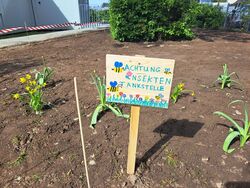 Insektentankstelle Das Bild zeit ein Beet auf dem frisch gepflanzte Blumen und ein Schild zu sehen sind. Auf dem Schild steht: "Achtung Insekten Tankstelle"