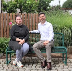 Auf dem Bild zu sehen sind Marina Drewes und Bürgermeister Jochen Engel sitzend auf einer grünen Bank vorm Rathaus Trebur