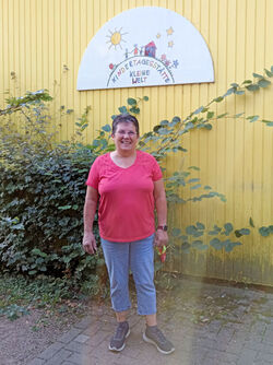 Die "Chefin" vor dem alten Gebäude Auf dem Foto ist Monika Kraft vor dem Gebäude des alten Kindergartens zu sehen. Über Ihr hängt ein Schild an der Wand auf dem in der Art einer Kinderzeichnung eine Sonne, Kinder und ein Haus zu sehen sind. Darunter steht: "Kindertagesstätte Kleine Welt"