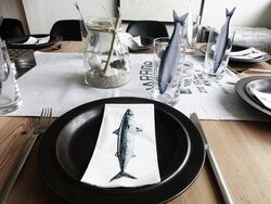 Auf dem Bild ist ein eingedeckter Tisch zu sehen und auf den Tellern liegen Servierten mit einem Hering drauf