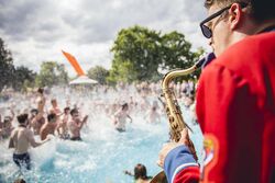 Das Bild zeigt einen Saxophonist am Beckenrand des Freibads und Tanze Fans im Wasser