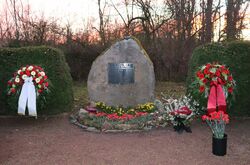 Das Bild zeigt den Gedenkstein mit Blumenkränzen und mehrere Blumengestecke